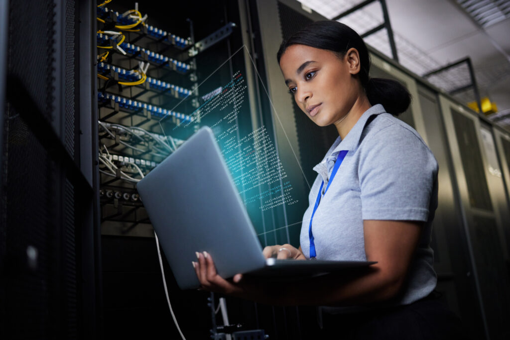 IT expert analyzing data on a laptop while standing next to stacks of data servers.