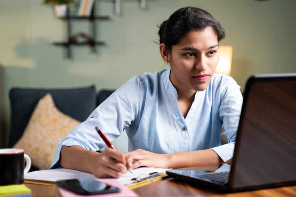 Young business woman writing down notes by looking laptop - concept of employee or student online training class at home