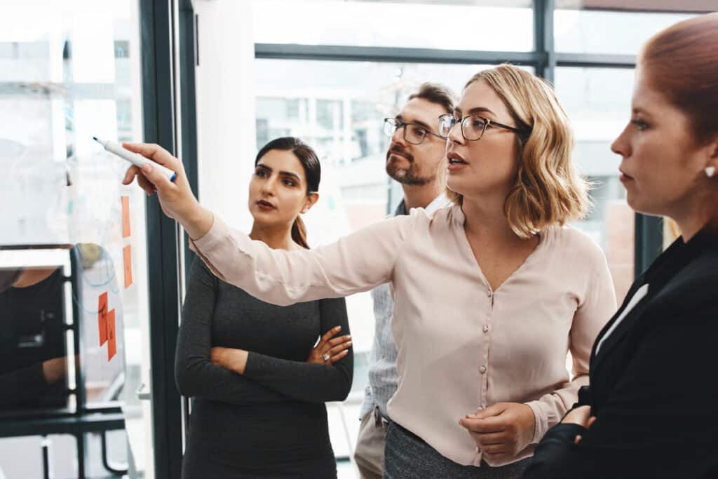 Woman, group and collaboration with notes on board with review, listening and planning with brainstorming. Coach, training and team by glass wall, questions and discussion with strategy in office