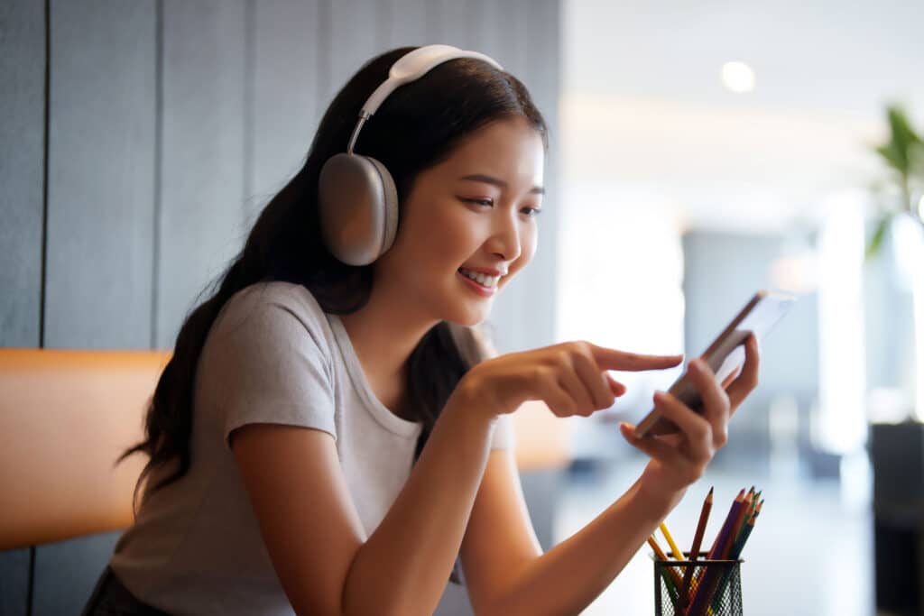 Asian teenage student woman wearing headphone to listening music and chatting with friends on smartphone for relaxation after studying and learning education knowledge for preparing test exam in cafe