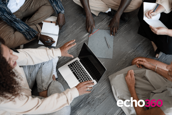 Adult learners sitting in a circle, with one holding a laptop towards the middle. Smiling and conversation can be seen by bodies apparently in motion.