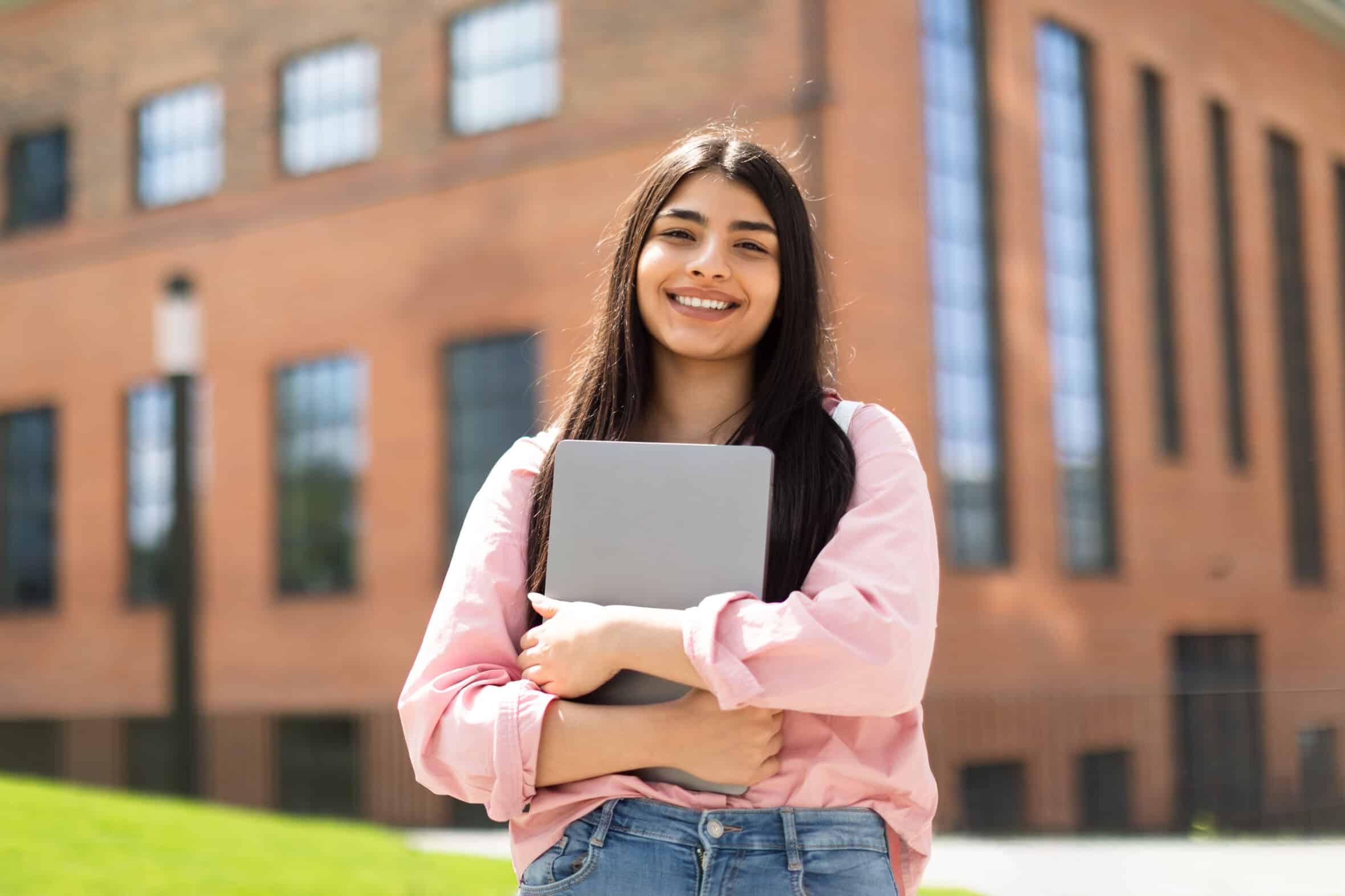 Portrait of happy hispanic lady student posing with laptop in hands outdoors, looking and smiling at camera