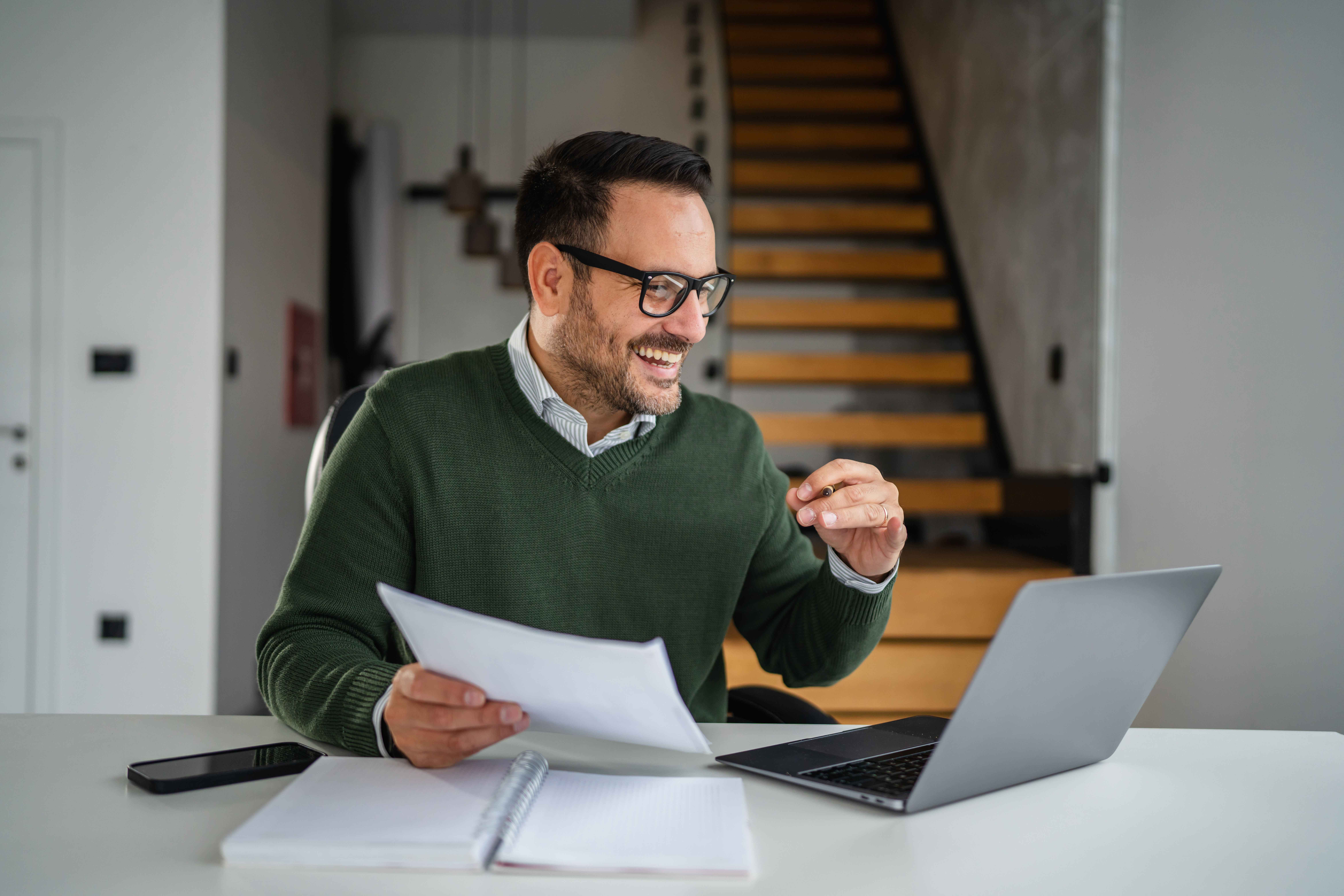 Happy businessman wearing glasses sitting at desk and actively participating in a video call or online meeting using a laptop in a modern home office setting
