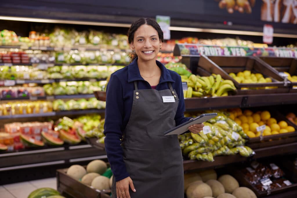 Smiling store employee standing near fruit display with digital tablet in hand. Young multiethnic woman in uniform posing confidently in fresh produce section of supermarket. Grocery staff member holding tablet while woking in fruit and vegetable area.