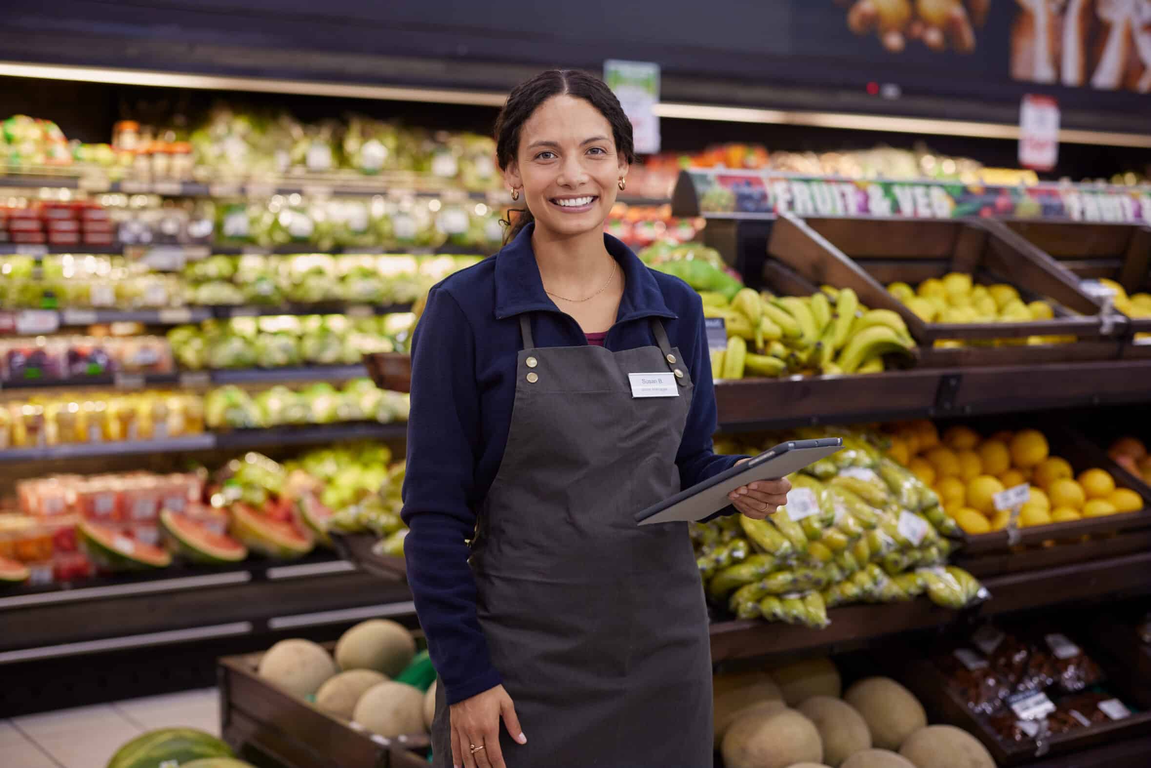 Smiling store employee standing near fruit display with digital tablet in hand. Young multiethnic woman in uniform posing confidently in fresh produce section of supermarket. Grocery staff member holding tablet while woking in fruit and vegetable area.